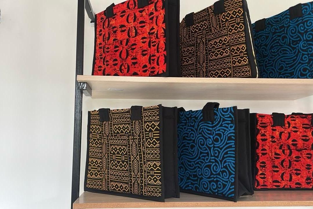 Kitenge tote bags in red, gold and blue prints, lined up on pale wooden shelves.